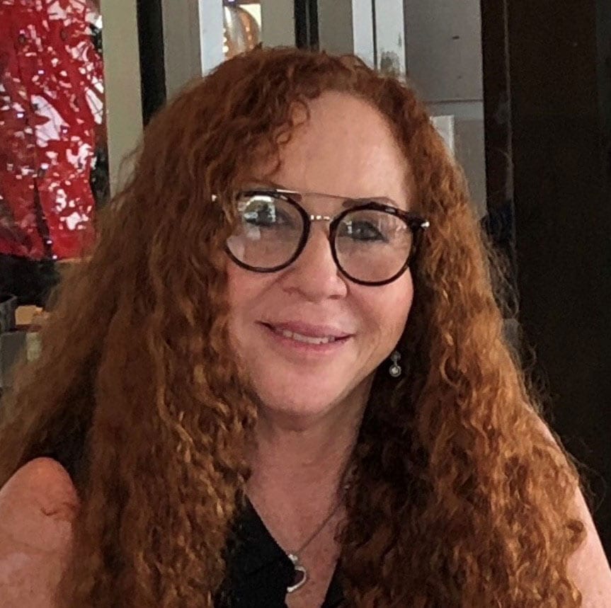 a smiling photo of Anny Smulevich, Board Member, a woman with long curly red hair and glasses smiles warmly. The background is a softly blurred indoor setting.