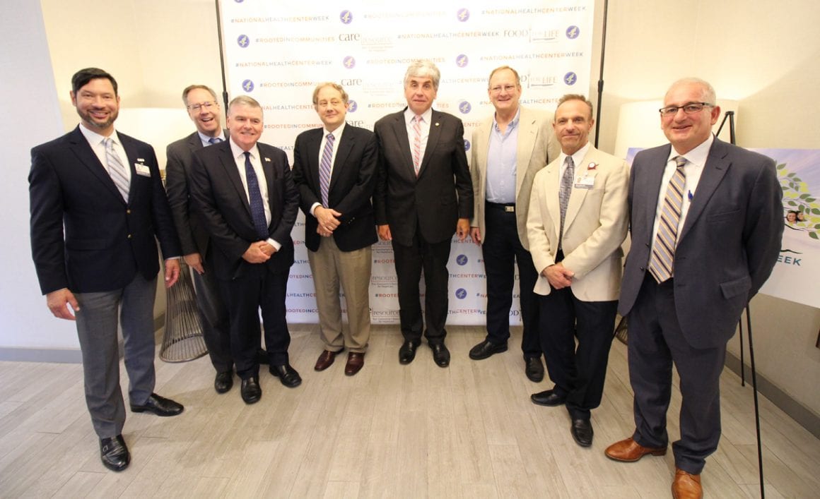 A group of eight men in suits and ties posing with HHS dignitaries in 2019 during their visit to Midtown Miami