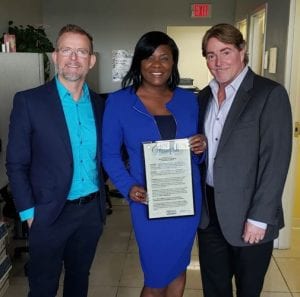 Three people stand smiling in an office. The woman in the center holds a framed certificate, wearing a bright blue suit. The tone is celebratory.