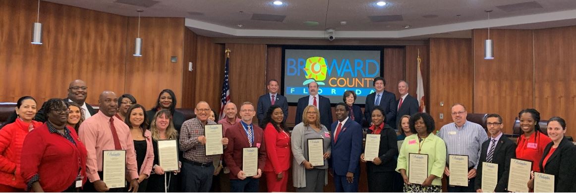 A diverse group of smiling individuals stands in a formal room holding certificates. The backdrop displays Broward County Florida. The mood is celebratory.