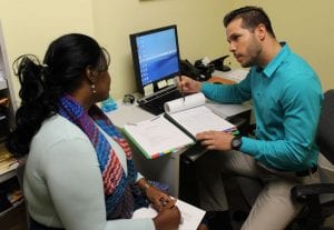 Photo of an intake process at Care Resource shows a man in a teal shirt consulting with a woman in a colorful scarf at a desk with a computer and documents, conveying a professional setting.