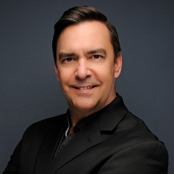 Photo of Daniel Falcon, a man in a black suit with a confident smile stands against a dark gray background. His arms are crossed, creating a professional and approachable demeanor.