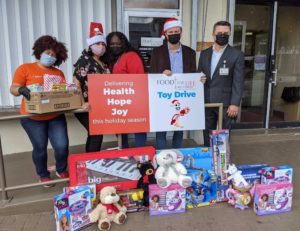 A group of people in festive attire hold a Food for Life Toy Drive sign, surrounded by colorful boxed toys and plush animals. They are smiling and wearing masks.