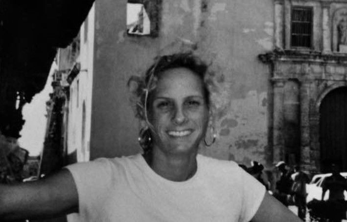 Beatriz Maria de la Sierra smiling with short hair in a white shirt, standing in front of an old building with textured walls, exuding a relaxed and cheerful vibe.