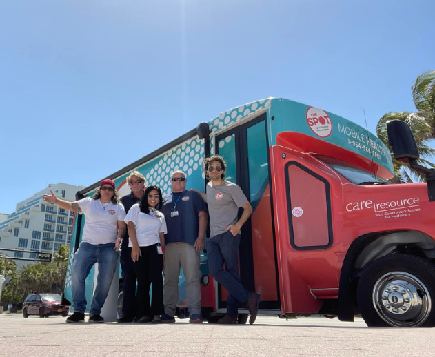 A group of five people stands smiling in front of The SPOT in Fort Lauderdale, a colorful mobile health clinic vehicle under a clear blue sky, conveying a cheerful, community-focused atmosphere.