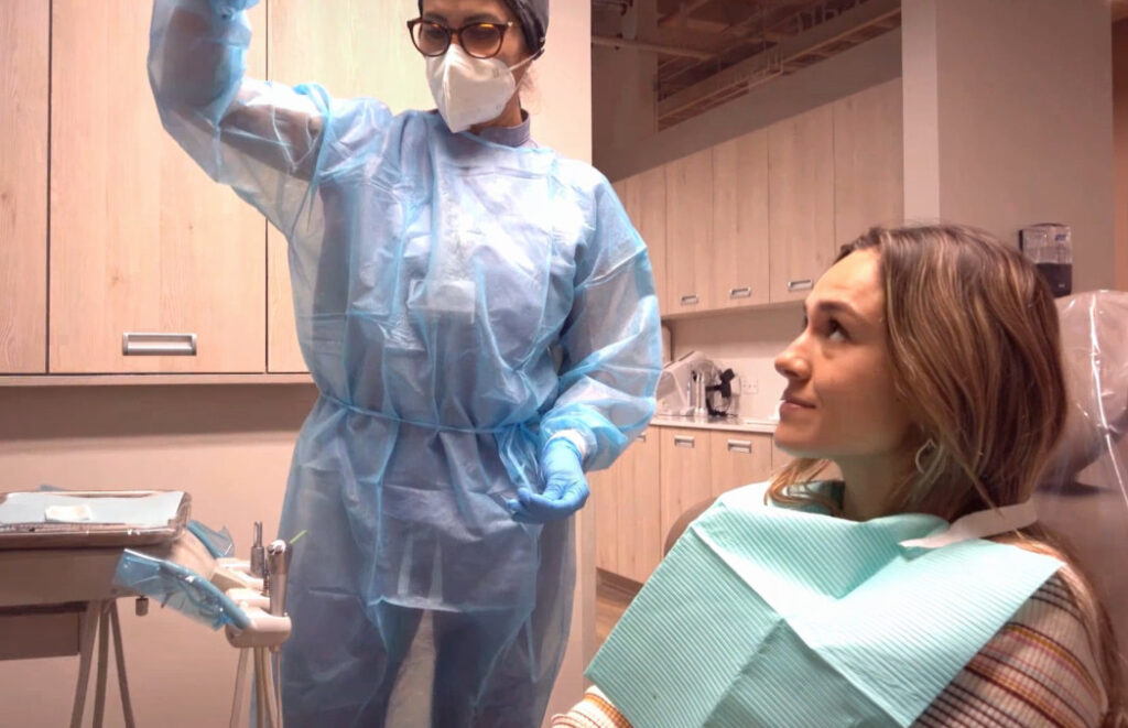 A dentist in protective gear, including a mask and gown, stands beside a seated patient in a dental office. The patient looks up, appearing calm.