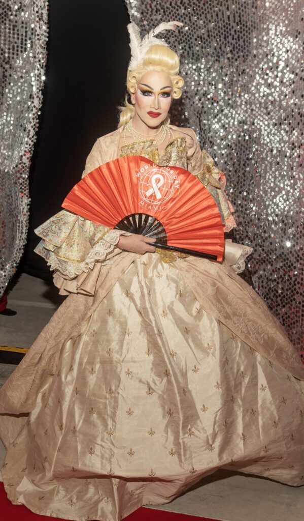 A person in an ornate, vintage-style gown holds a red fan with a logo, standing against shimmering silver curtains. The tone is elegant and theatrical.