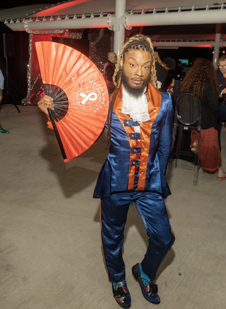 Man in vivid blue and orange suit poses confidently, holding a large red fan. The background shows a stylish event with seated guests.