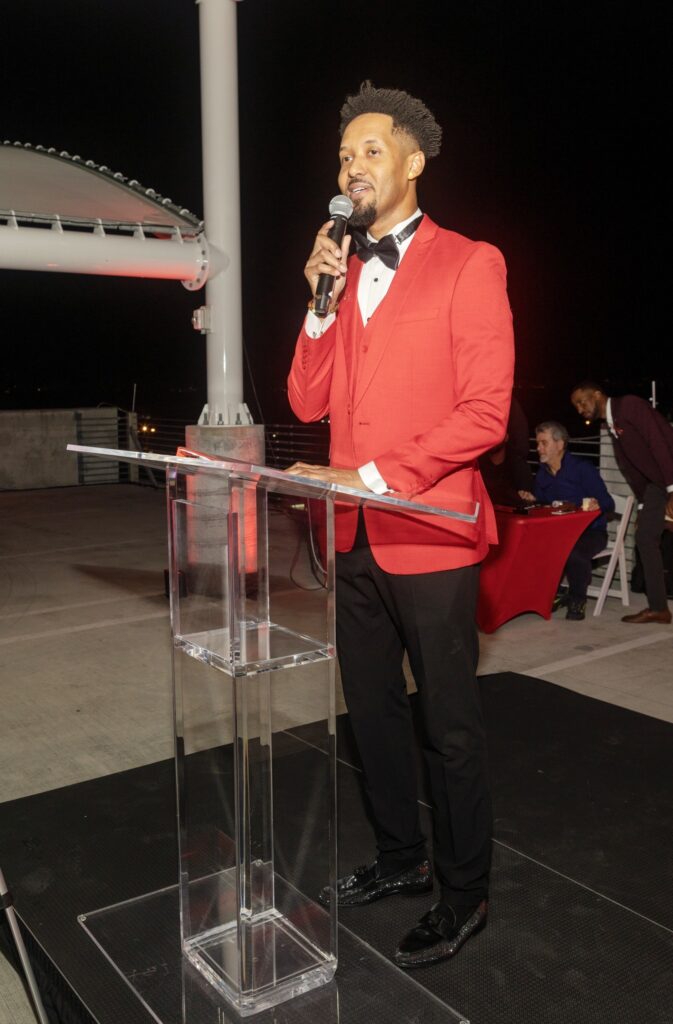 Man in a bright red tuxedo, speaking confidently at a podium with a microphone. Nighttime rooftop setting, elegant and formal atmosphere.
