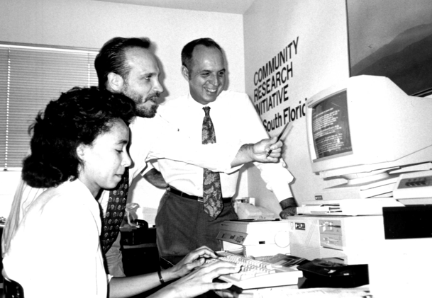 A black and white photo of three people in an office working at a vintage computer. A sign on the wall reads Community Research Initiative, South Florida.