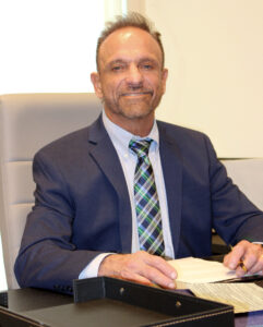 For Project HIMPACT, A man in a suit sits at a desk, smiling confidently. He holds a pen over documents, with a neat stack of papers in front. Bright, professional setting.