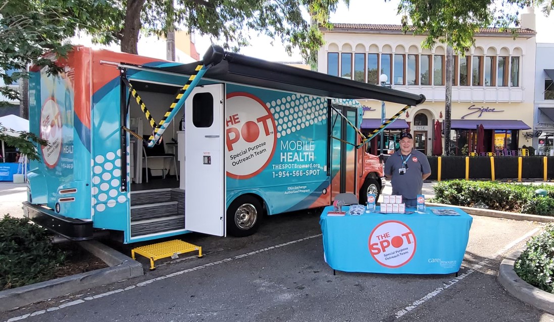 Mobile health van with The SPOT branding parked on a street, offering health services. A staff member stands beside a table with informational materials.