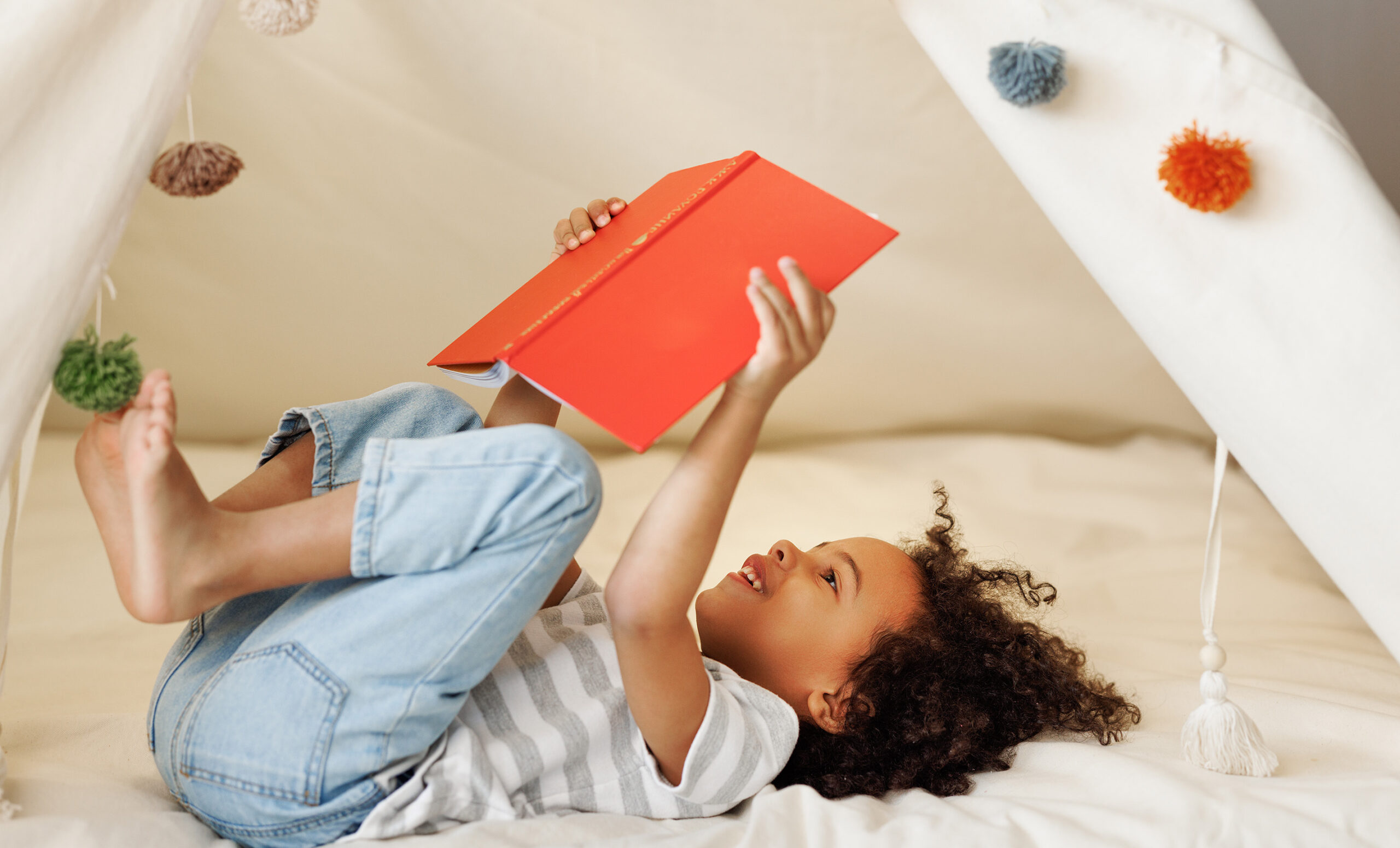 Child lying in a cozy tent, holding an open red book and smiling, with soft cushions and hanging pom-poms—playful imagery for pediatrics storytelling.