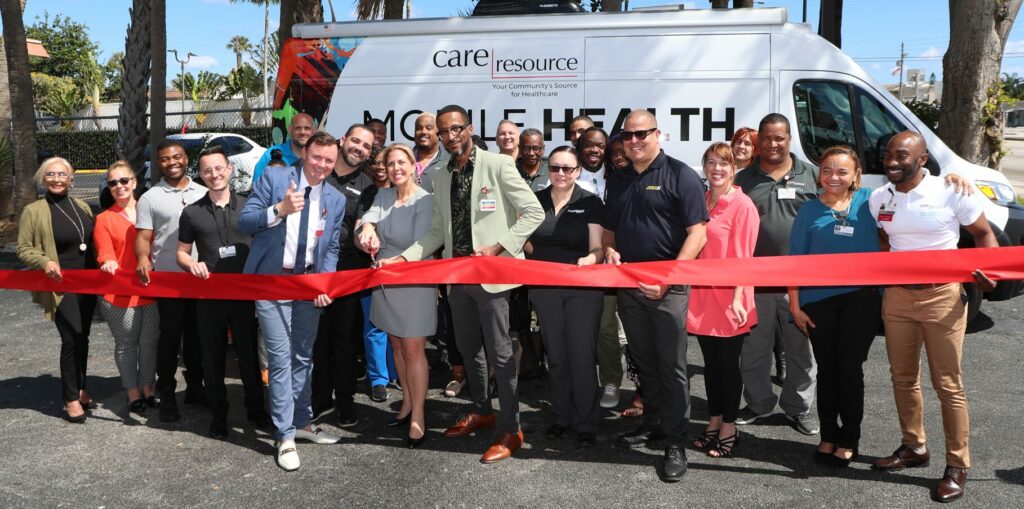 A diverse group of people smiles and holds a red ribbon in front of a mobile health van dedicated by Advance Auto Parts Foundation, for a ribbon-cutting ceremony.