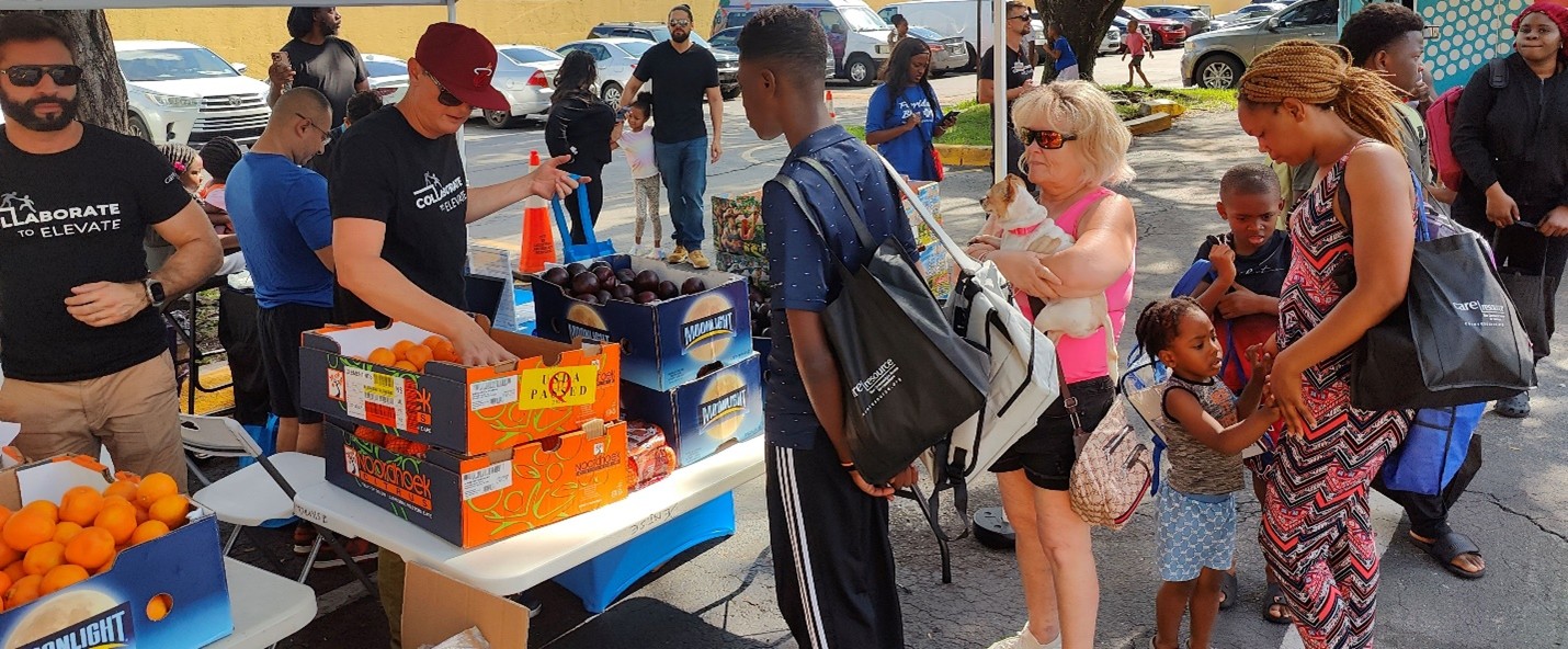 A diverse group of people gathers at an outdoor food distribution. Volunteers offer fresh produce like oranges and avocados. The atmosphere is lively and communal.