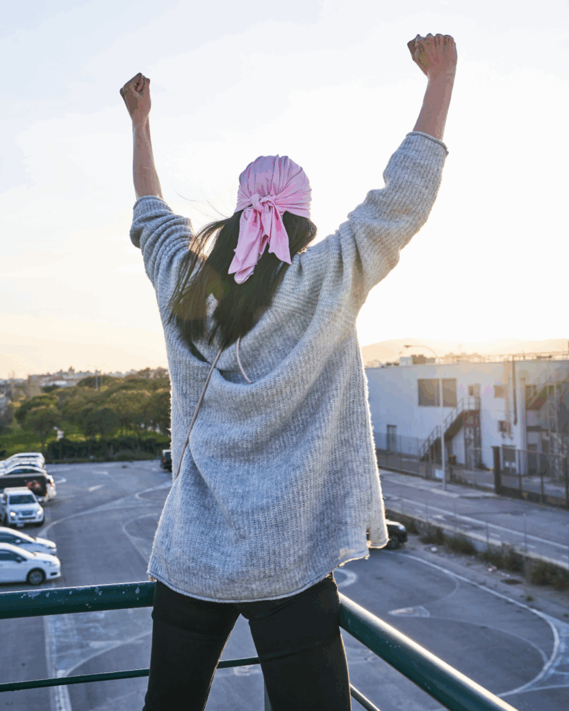 A person stands triumphantly with raised arms, wearing a pink headscarf and gray sweater, overlooking a parking lot at sunset. The scene conveys joy and victory.