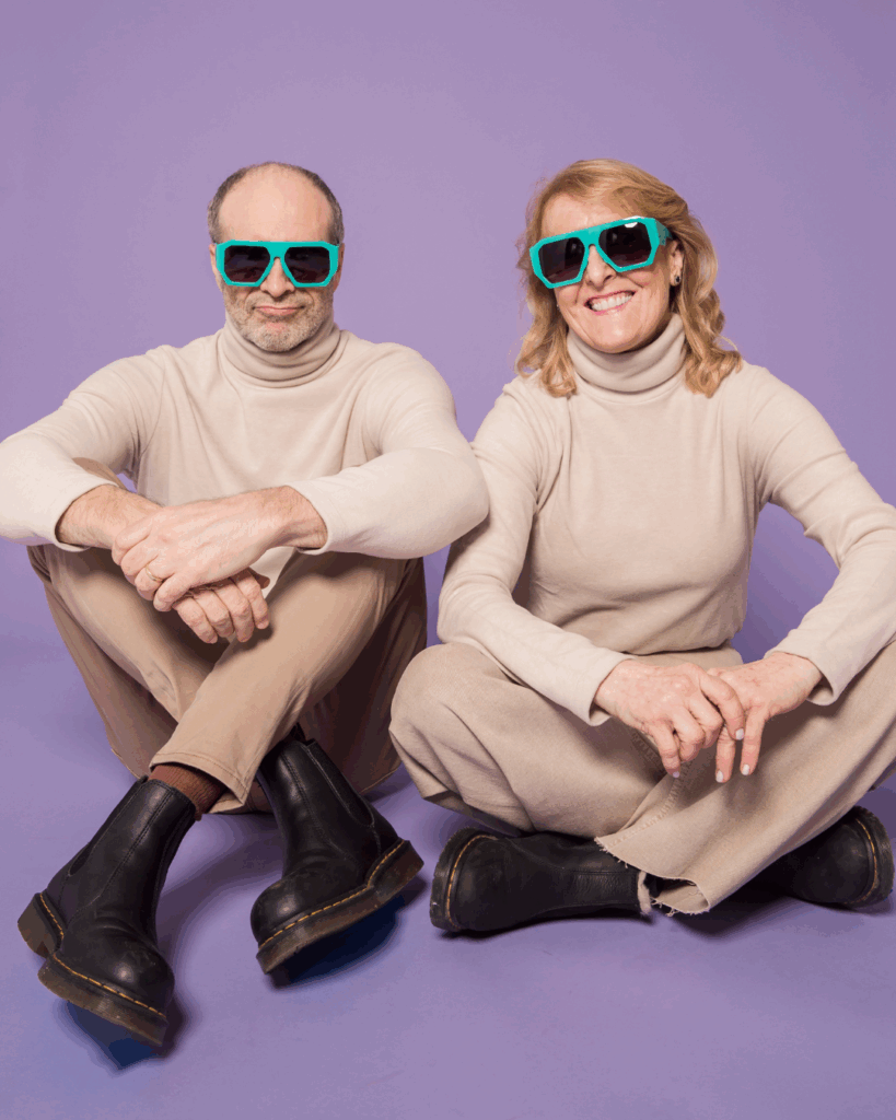 A man and woman sit cross-legged on a purple floor, wearing matching beige outfits and large teal sunglasses, smiling warmly. Retro, playful vibe.
