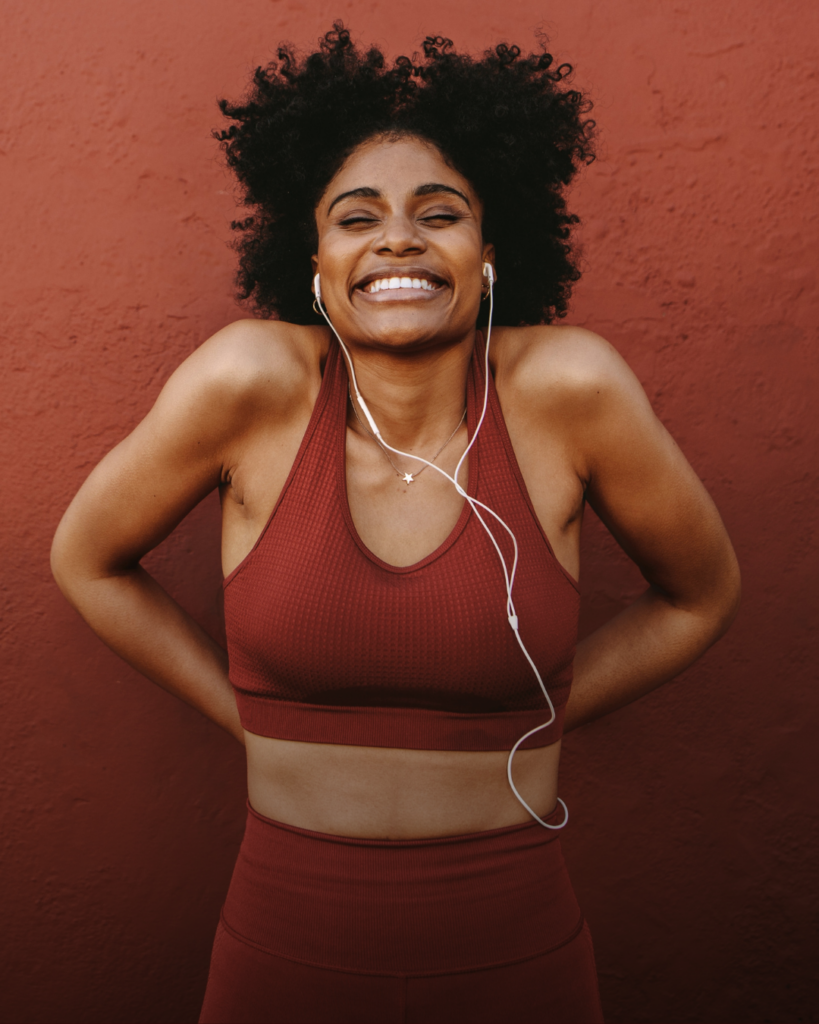 In awareness for Cervical health, a photo of a smiling woman with curly hair in a red sports outfit listens to music with earbuds against a textured red wall, radiating happiness and energy.