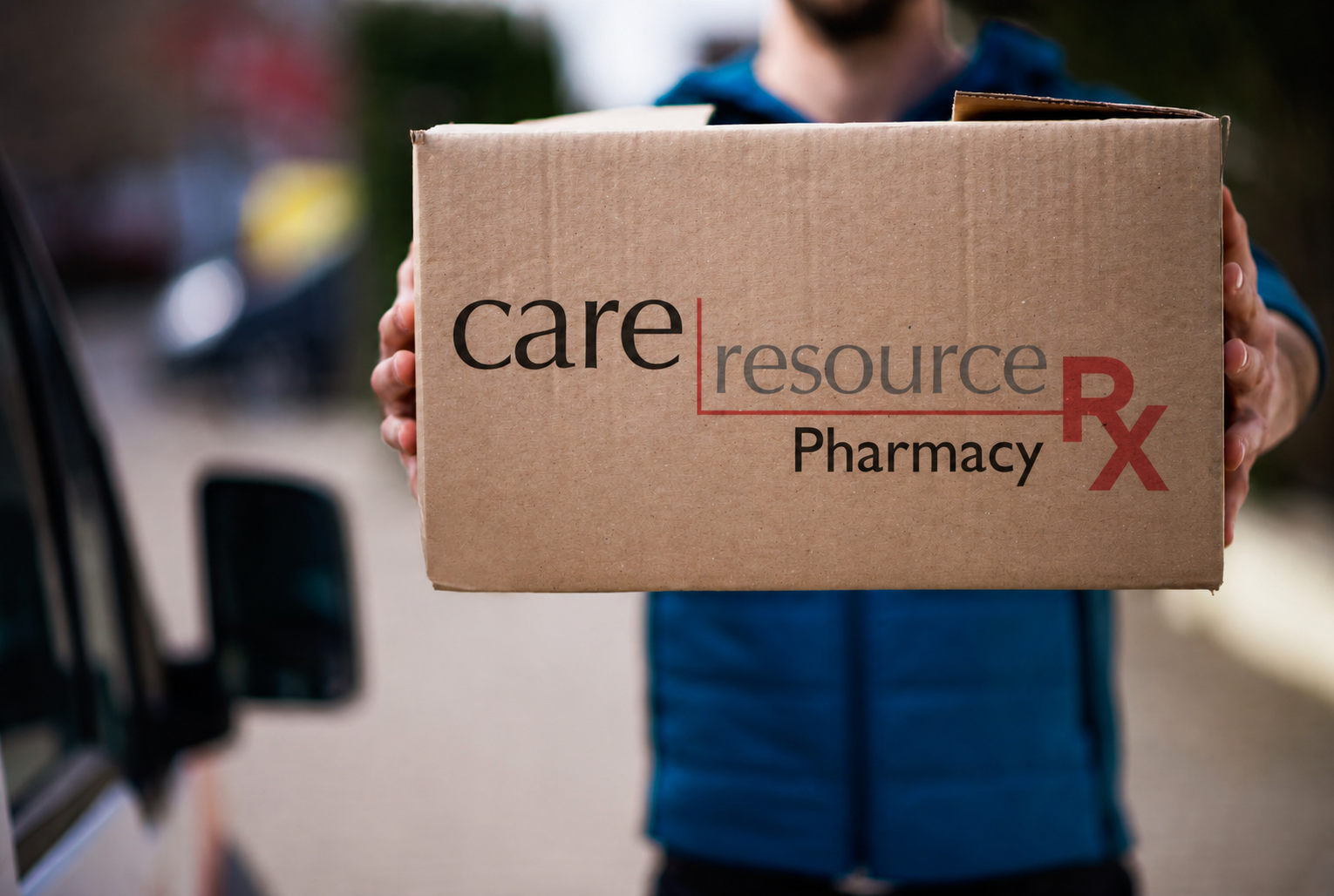 Delivery person holds a cardboard box labeled “care resource Pharmacy Rx” beside a vehicle, conveying accessible prescription medication delivery.