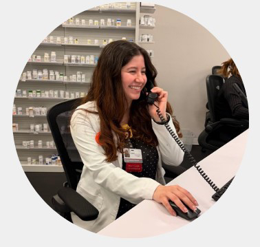 Smiling female pharmacist in a white coat speaks on a phone at a pharmacy counter, with medicine shelves behind her, conveying warmth and professionalism.