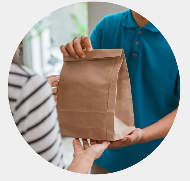 Two people exchange a brown paper delivery bag, suggesting contactless grocery or food delivery; hands and torsos visible in a bright setting.