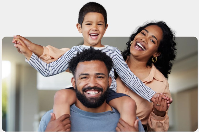 Smiling family at home: a young boy rides on his father's shoulders while his mother holds his hand, creating a joyful, loving scene.