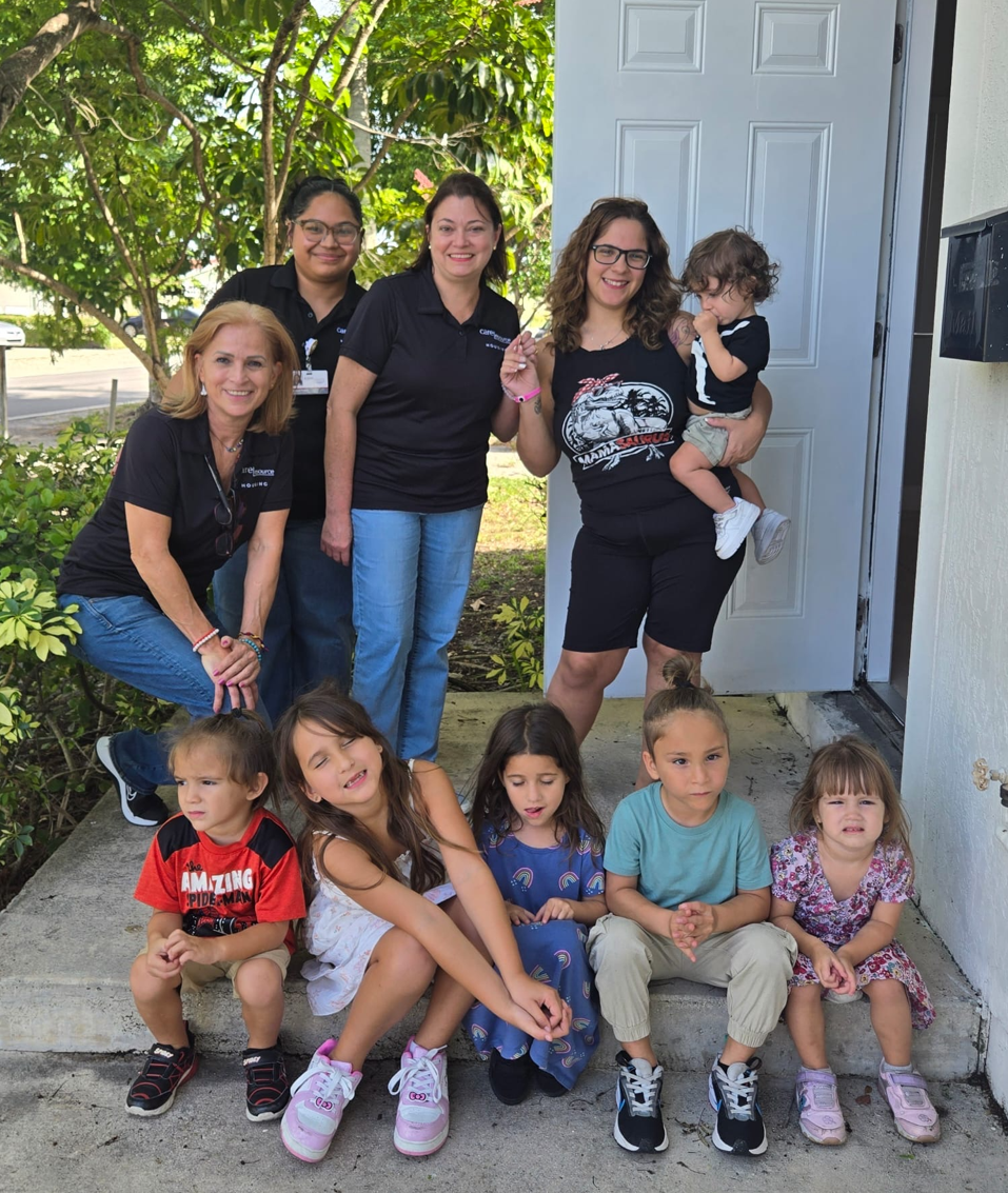 Four smiling women and six children pose outside a home, surrounded by greenery, creating a warm, cheerful family scene.